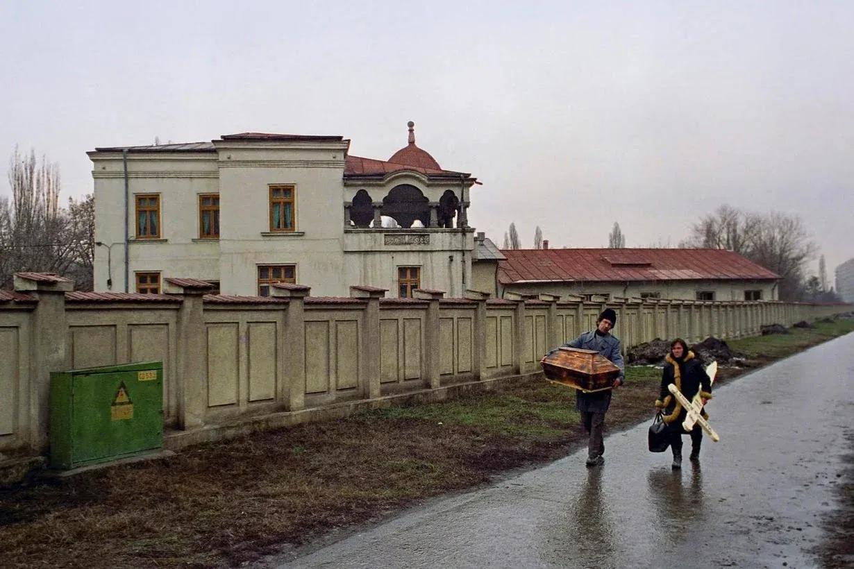 A man carrying a small wooden coffin and a woman carrying a white cross walk along a wet road beside a long wall